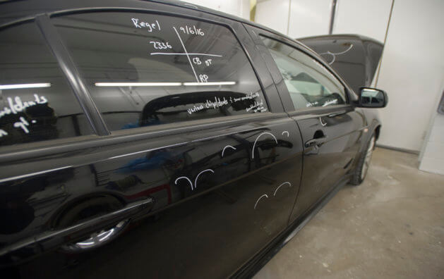 Photo of the side of a black car with a text written with white marker in the right window behind passengers, and stripes with the same marker on the doors.