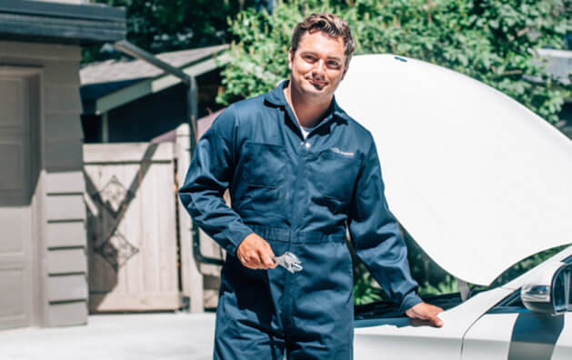 A man wearing workshop clothes holding a spanner in his hand, standing in front of a car with the hood open.