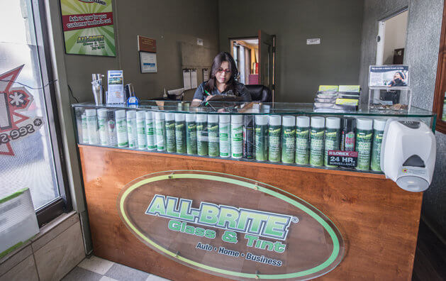 A woman standing behind a wooden and glass counter with the All-Brite Glass & Tint logo.